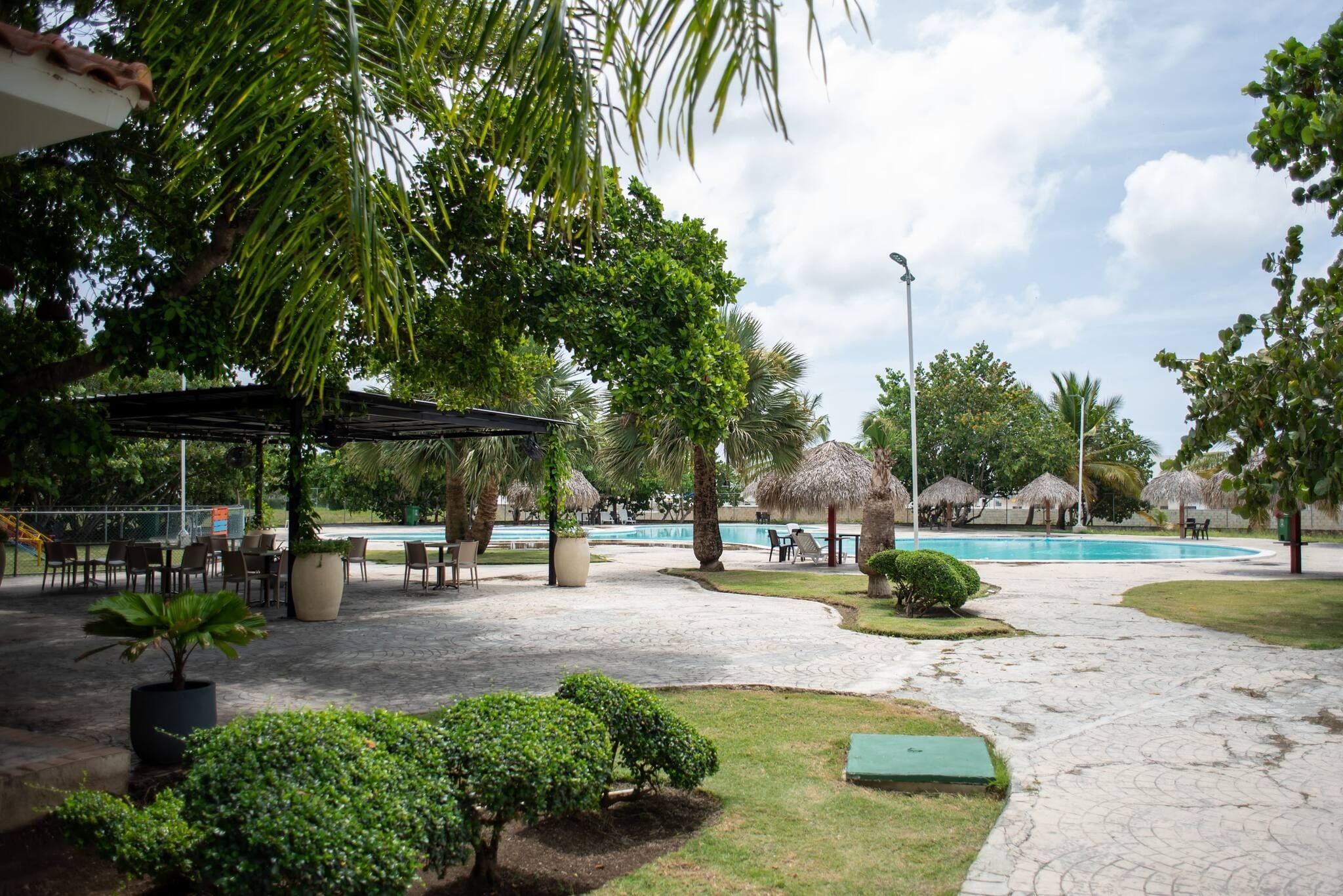 Zona de piscina con un área de estar cubierta, palmeras y una piscina azul cristalina.