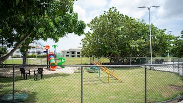 A fenced playground with various play equipment, including a slide and swings, surrounded by trees and a building in the background.
