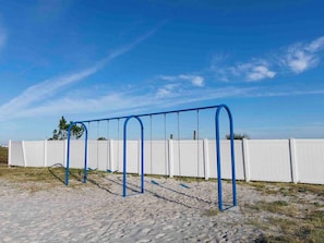 Um parquinho com balanço em área de areia, cercado por uma grade branca e céu azul claro.