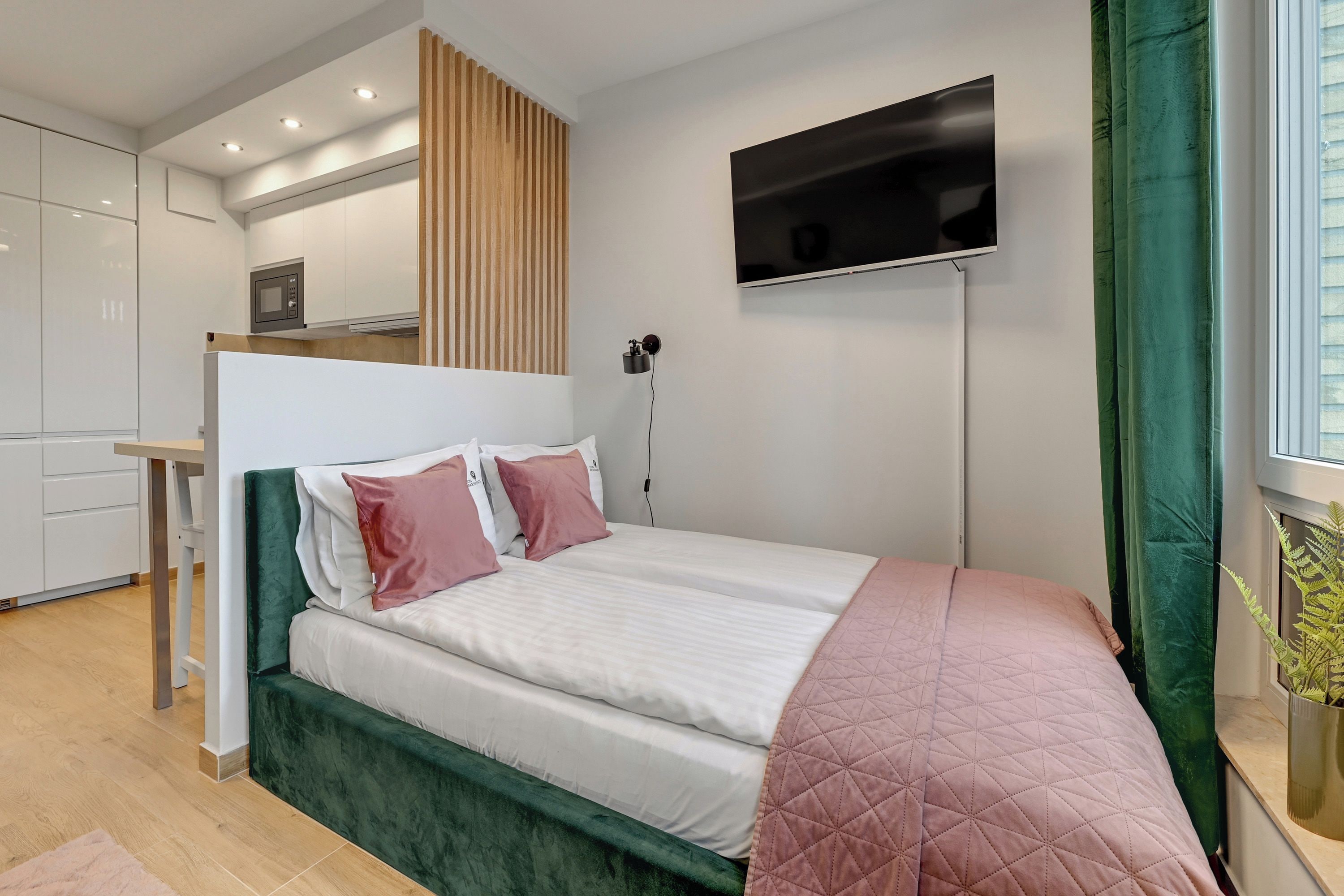 Une chambre moderne avec un lit, un téléviseur fixé au mur et un coin cuisine visible à travers une porte ouverte.