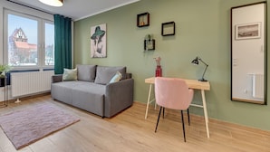 A modern living room with a grey sofa, a small desk with a pink chair, a radiator, and a window with curtains. - Studio Wilcza with Parking  (Gdańsk)