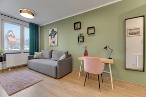 A modern living room with a grey sofa, a small desk with a pink chair, a radiator, and a window with curtains. - Studio Wilcza with Parking  (Gdańsk)