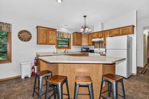 A kitchen with wooden cabinets, a central island with stools, a refrigerator, and a clock on the wall. - Stepaway Vacation Rentals/Hillcrest Cottages-Cedar Lodge (Saint Germain)
