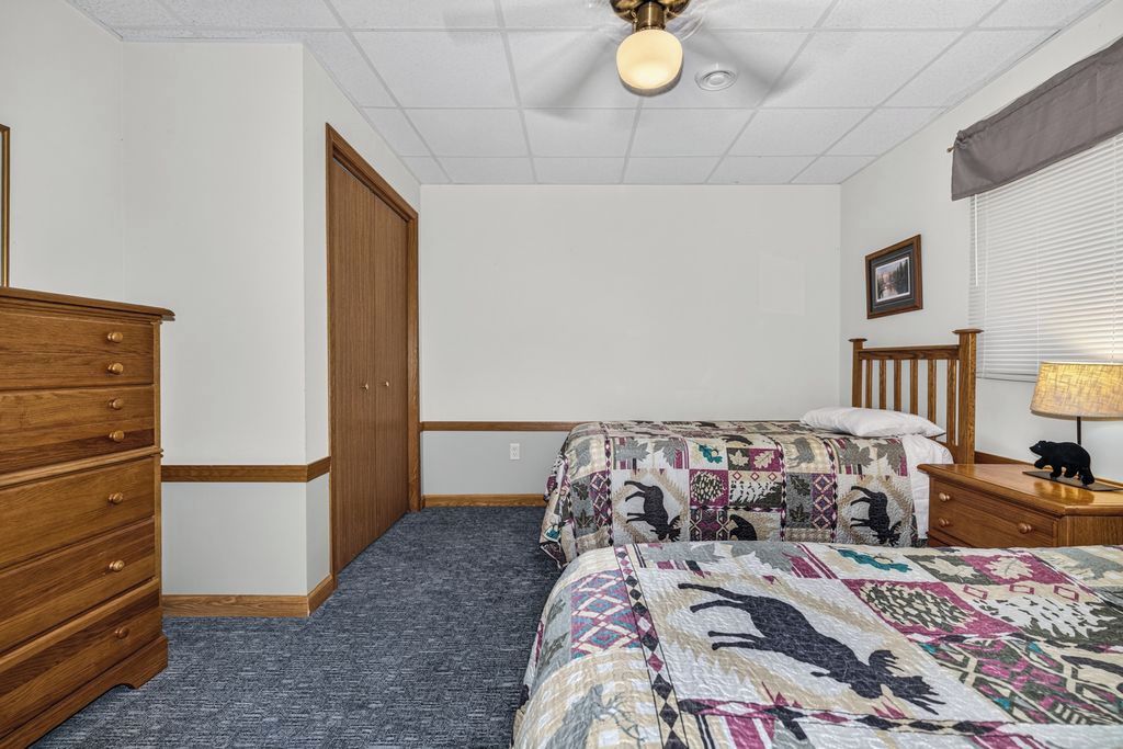 Ein Schlafzimmer mit einem Bett, einer Kommode, einem Nachttisch, einer Lampe, einem Deckenventilator und einem Fenster mit Jalousien.