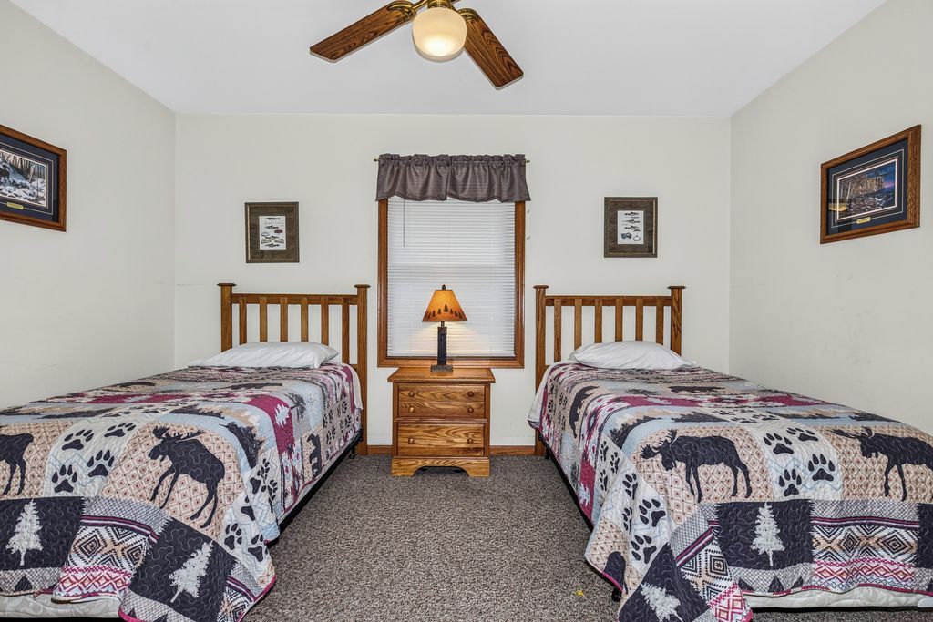 A bedroom with two single beds, a nightstand with a lamp, a ceiling fan, and framed pictures on the walls.