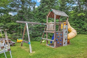 A wooden playground with a slide and climbing wall, surrounded by grass and trees. - Stepaway Vacation Rentals/Hillcrest Cottages-Maple (St.Germain)