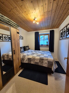 Ein Schlafzimmer mit Holzboden, einem Bett mit gemustertem Überwurf, einem Fenster mit Vorhängen und Wandgestaltungen.