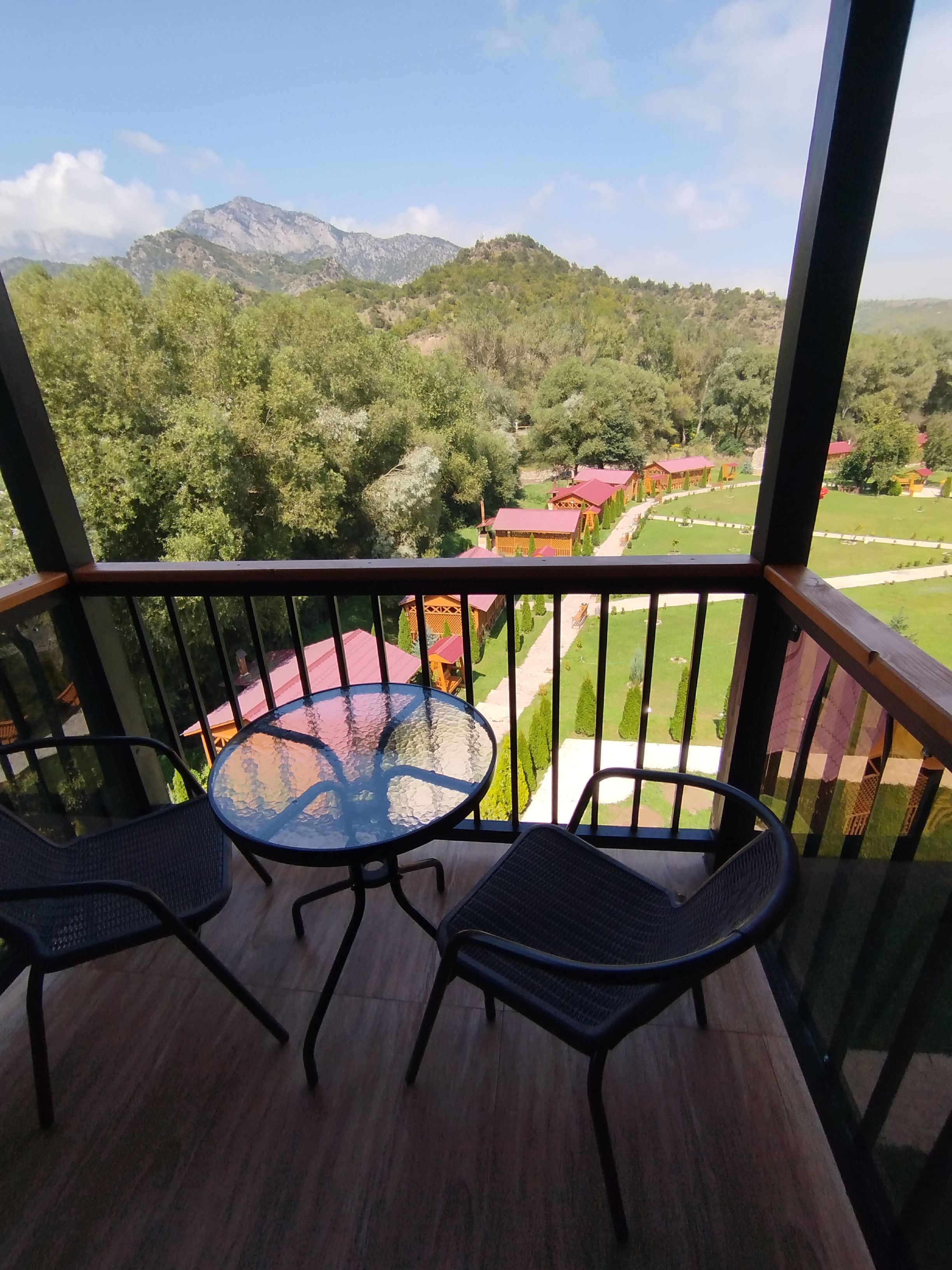 A balcony with a glass table and two chairs, overlooking a green landscape with buildings.