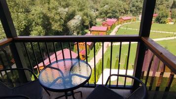 A balcony with a glass table and two chairs, overlooking a green landscape with buildings.