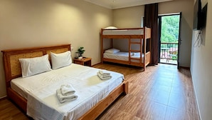 Quarto de hotel com uma cama grande, uma beliche menor, mesa de cabeceira com planta e varanda com mobiliário externo.