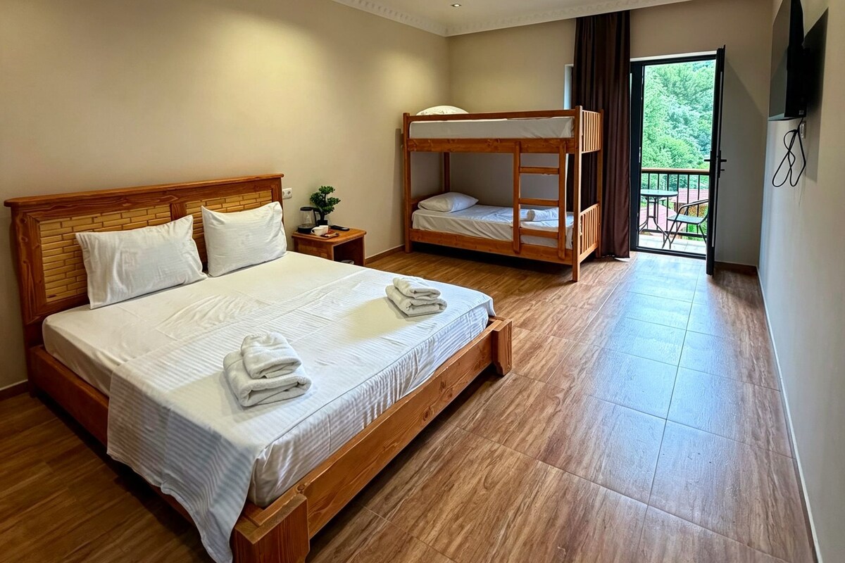 A hotel room with a large bed, a smaller bunk bed, a nightstand with a plant, and a balcony with outdoor furniture.