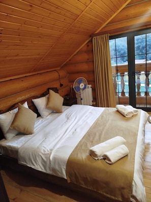 A wooden cabin bedroom with a bed, pillows, a fan, and a view through a window.