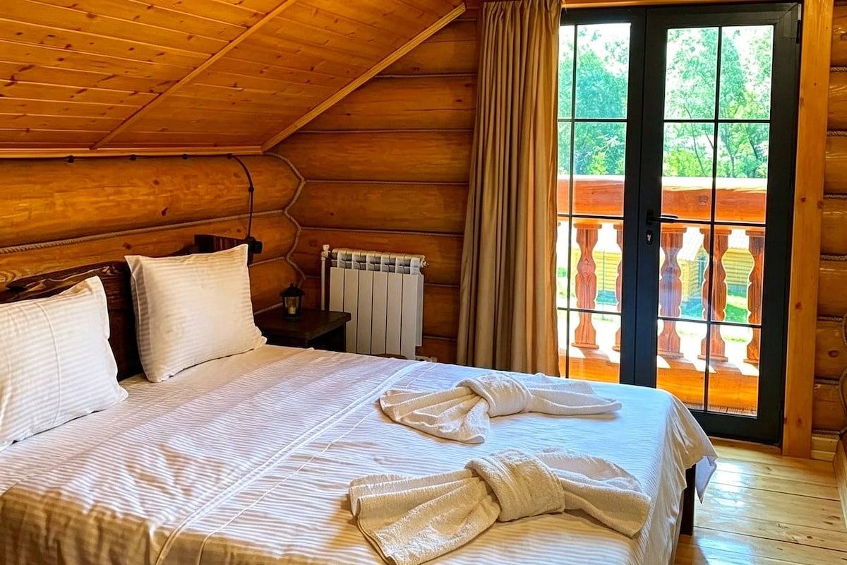A wooden cabin room with a bed, pillows, a nightstand, a lamp, a radiator, and a view of a balcony.
