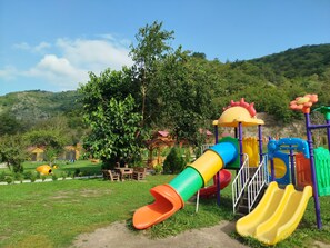 A playground with slides and climbing structures, surrounded by greenery and hills.