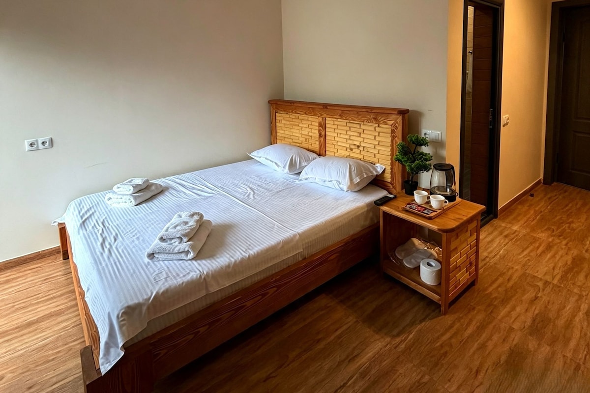 A neatly made bed with white linens and towels, a wooden headboard, a bedside table with a plant, and a nightstand with a kettle and cups.