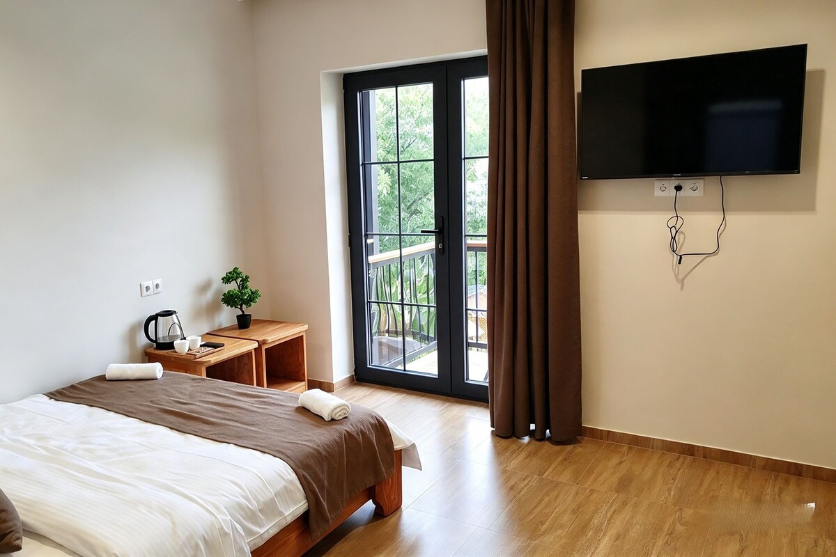 A hotel room with a bed, a small table with a kettle, cups, and a plant, a TV mounted on the wall, and a large window with curtains.