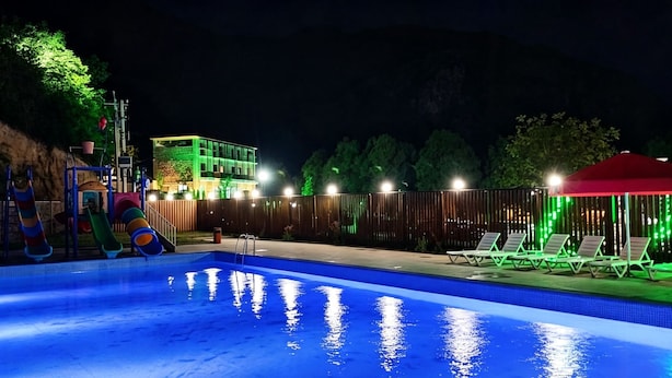 Piscina com escorregador, cercada por uma grade, e um edifício com iluminação verde ao fundo.