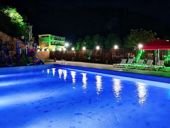 A swimming pool with a slide, surrounded by a fence, and a building with green lighting in the background. - Sonat Resort (Gosh)