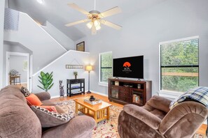 A modern living room with a flat-screen TV, a ceiling fan, and a staircase. - Hidden Creek Hideaway | Hot Tub, 2 Acres & Near Jacob's Well (Wimberley)