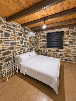 Ein Schlafzimmer mit einer Steinwand, einer Holzbalkendecke, einem Bett, einem Fenster mit Jalousien und einer Hängelampe.