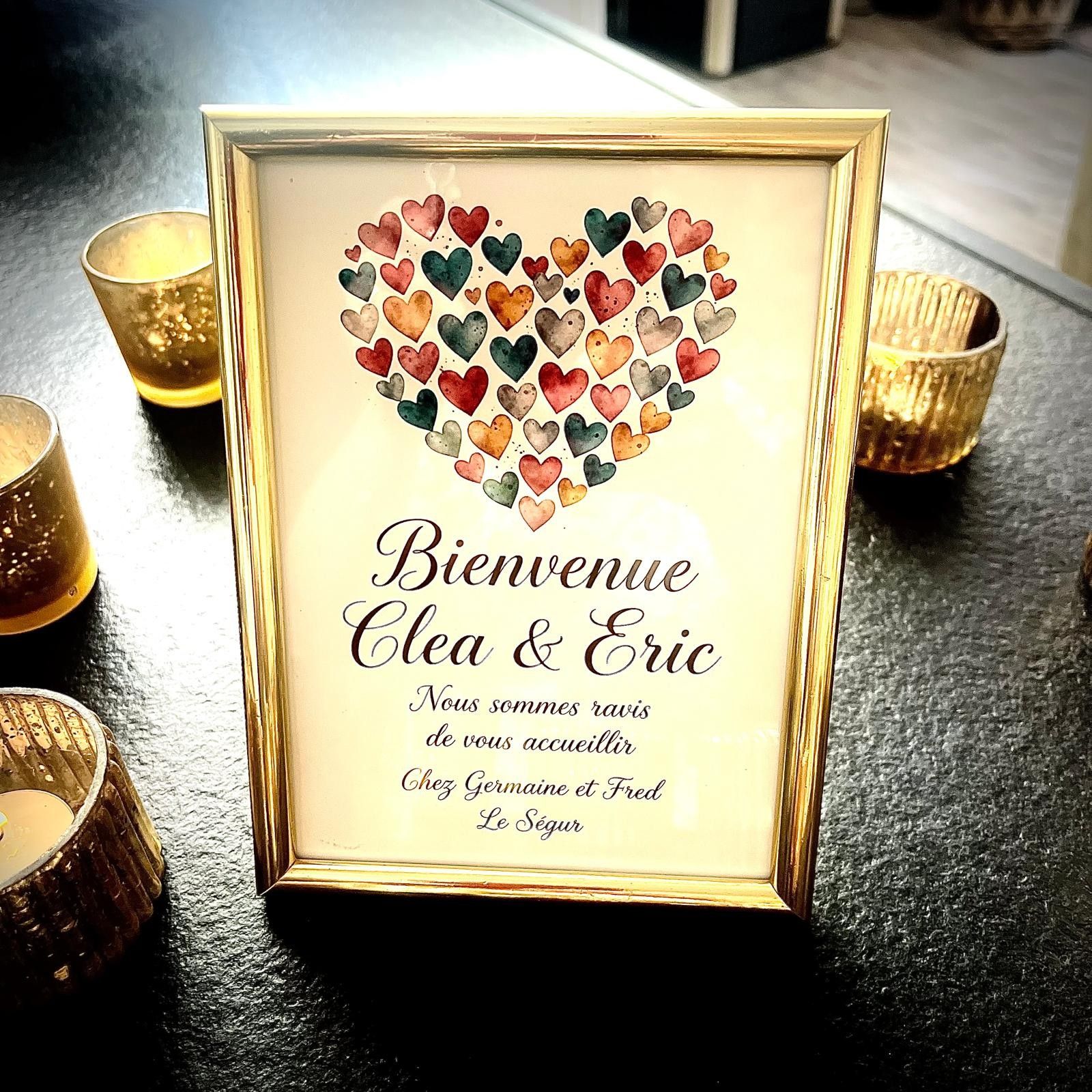 Une carte de bienvenue encadrée, ornée d’un collage en forme de cœur composé de cœurs de différentes couleurs, et portant le texte suivant : « Bienvenue, Cléa et Éric ».