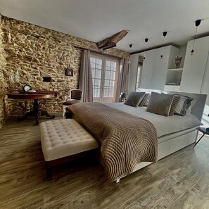 A bedroom with a stone wall, a bed with patterned bedding, a wooden bench, a small table with a clock, and a window with curtains.