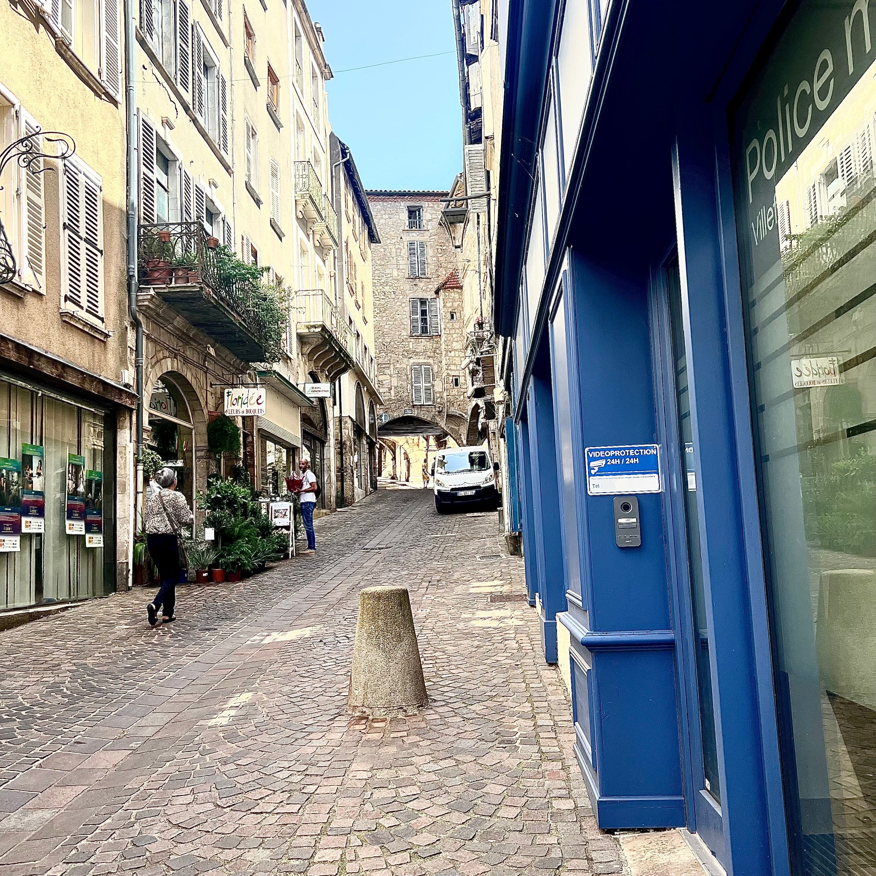 Una stretta strada acciottolata fiancheggiata da edifici, una stazione di polizia blu e una persona che cammina.