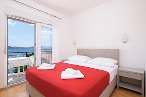 A bedroom with a red bedspread, white pillows, and a view of the sea through a sliding door. - A2 apartment with shared pool & balcony, next to the beach (Mastrinka)