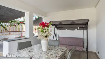 A room with a dining table, chairs, a white vase with flowers, a swing, and a view of a garden.