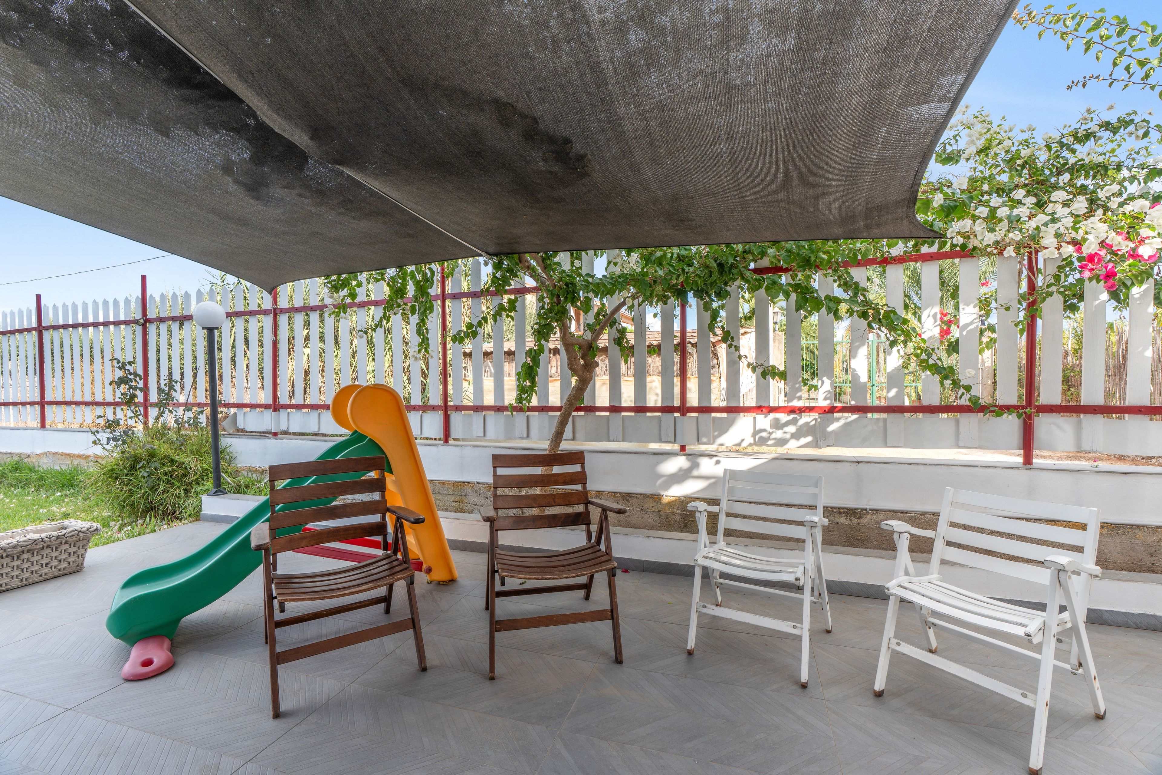 A covered outdoor seating area with a children's slide, benches, and a white picket fence.