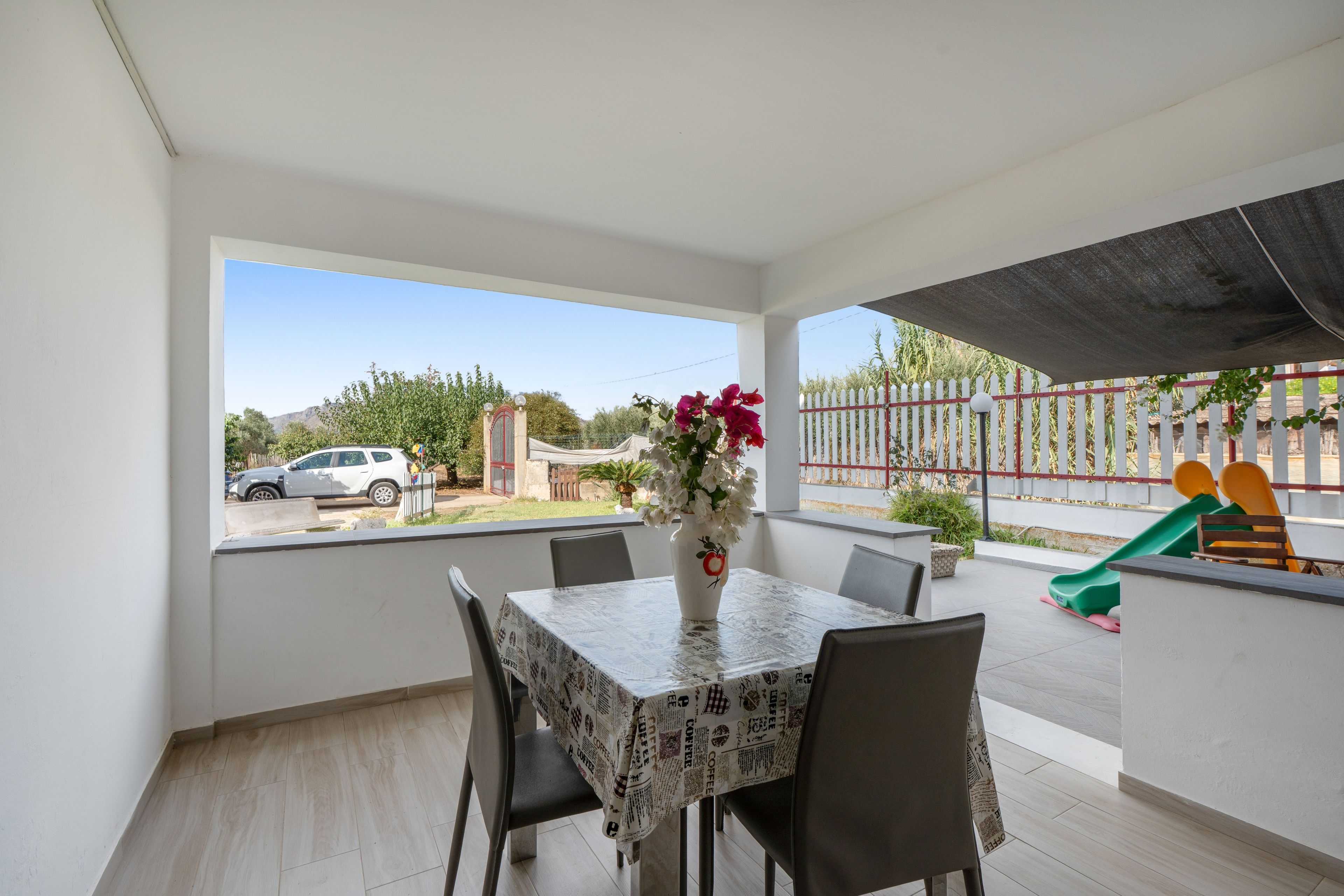 Ein Balkon mit Tisch und Stühlen, einer Blumenvase sowie Blick auf einen Spielplatz und ein parkendes Auto.