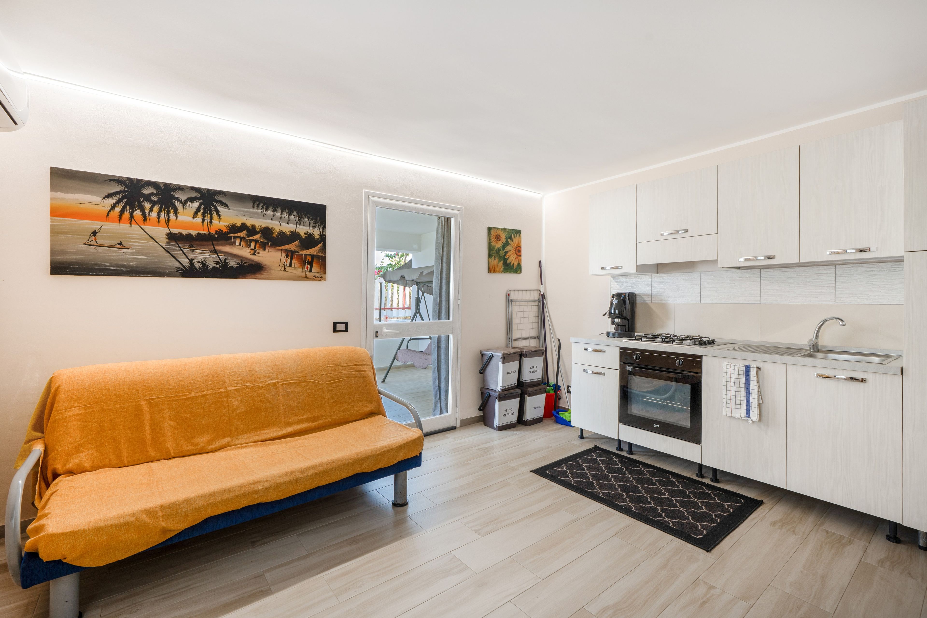 Eine moderne Küche mit weißen Schränken, einem Backofen und einer Spüle. Im Wohnbereich befindet sich eine gelbe Couch, und an der Wand hängt ein Gemälde von einer Strandansicht.