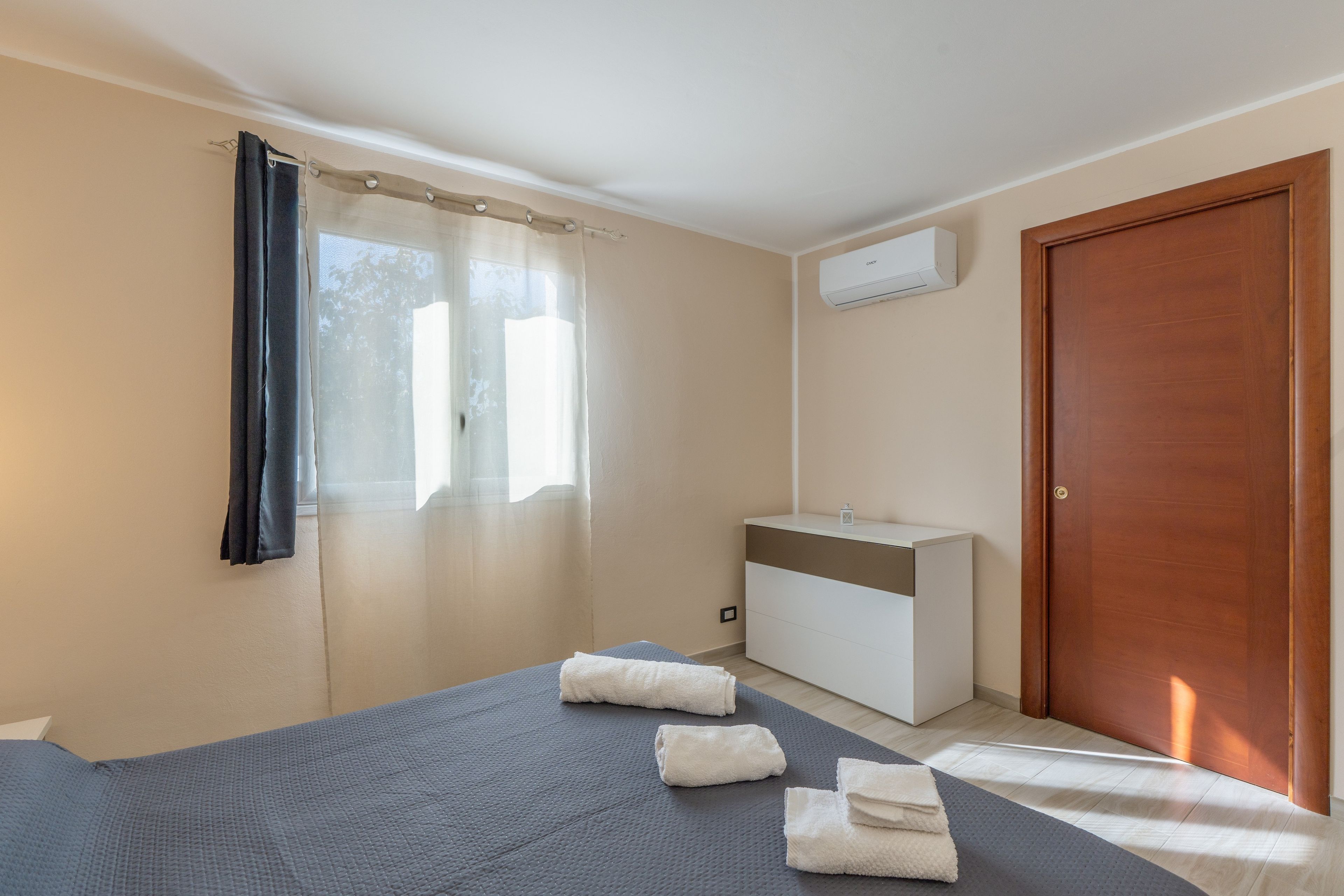 A bedroom with a bed, a bedside table, an air conditioning unit, a door, and a window with curtains.