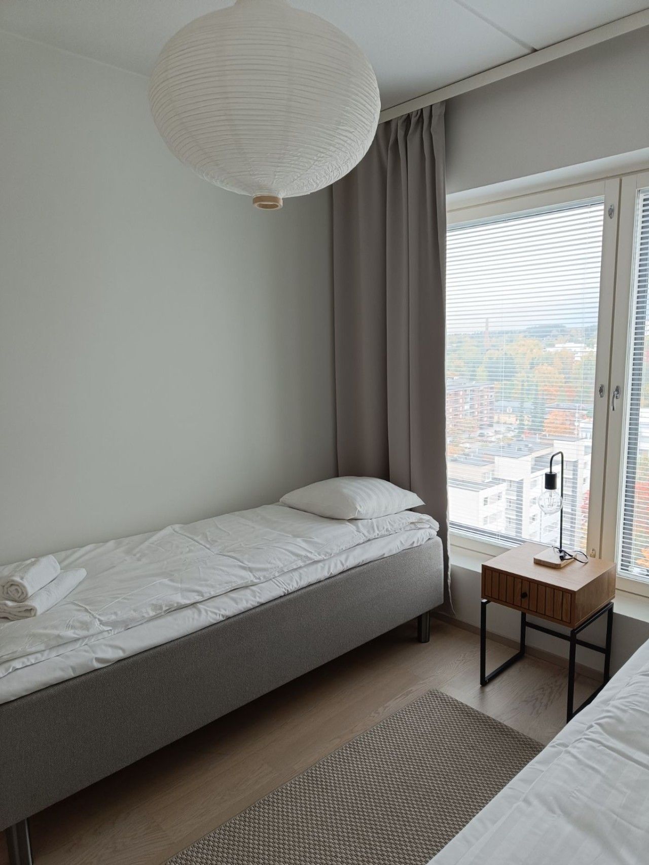 A single bed with a grey mattress and white bedding, a bedside table with a lamp, a window with blinds, and a view of buildings outside.