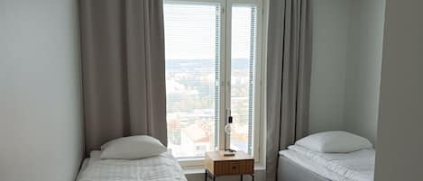 Un dormitorio moderno con una cama individual, una cama más pequeña, una mesita de noche, una lámpara y vistas de la ciudad a través de amplias ventanas.