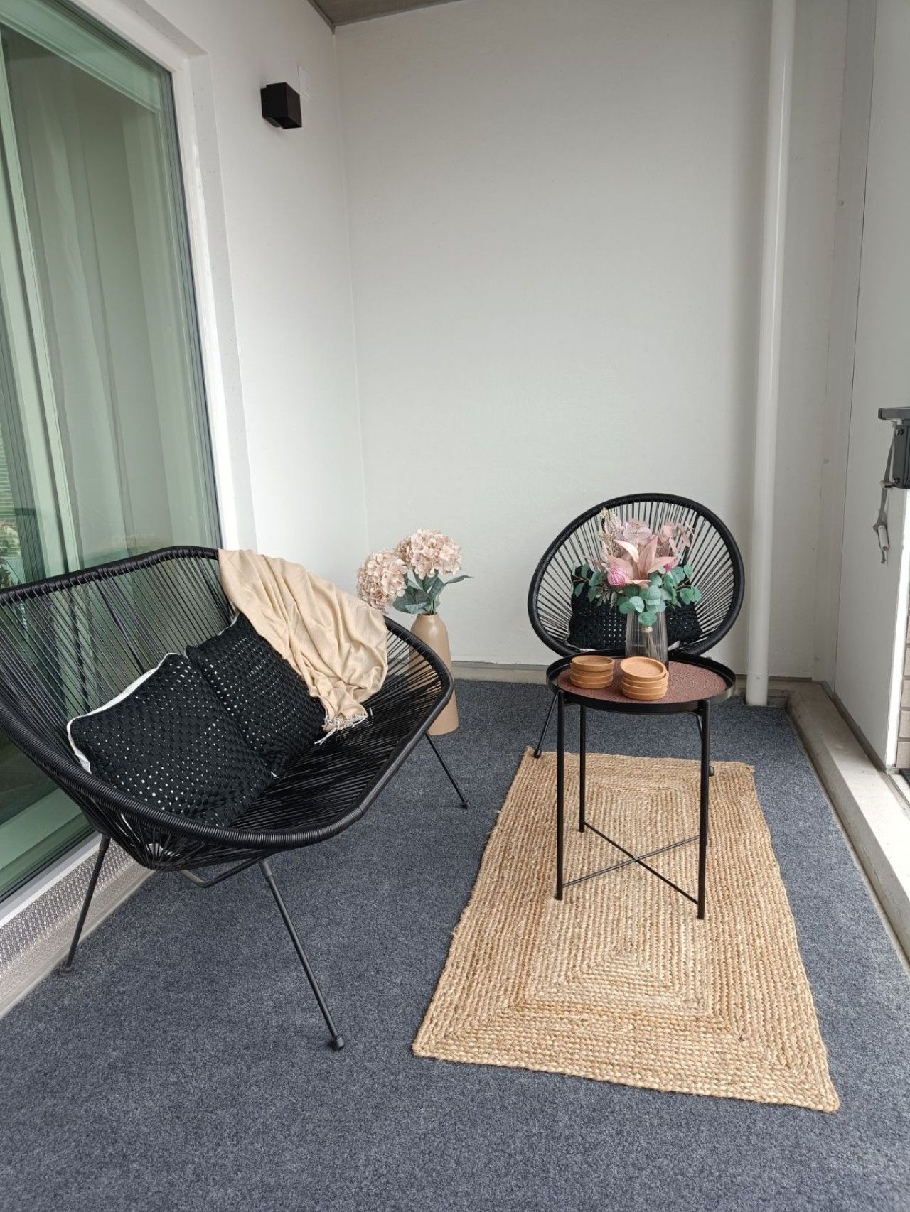Un pequeño balcón resguardado con una silla de mimbre negro, una mesa redonda con un florero y una alfombra tejida en el suelo.