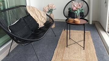Un pequeño balcón resguardado con una silla de mimbre negro, una mesa redonda con un florero y una alfombra tejida en el suelo.