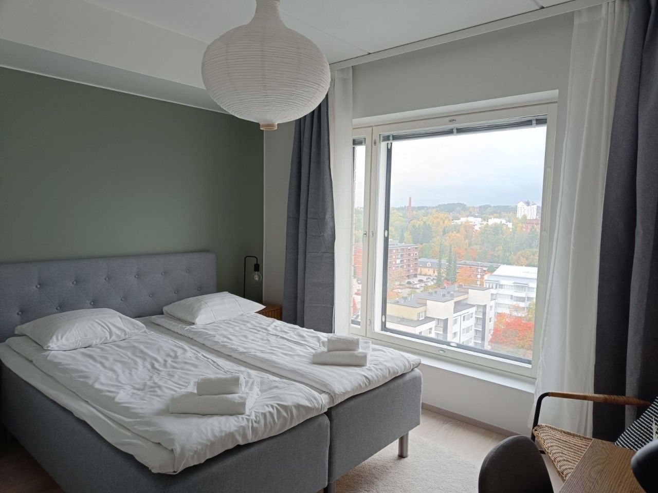 Un dormitorio con una cama grande, vistas de la ciudad a través de una ventana y una lámpara colgante.