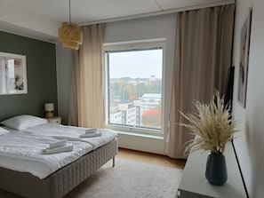 Una camera da letto con una grande finestra, un letto, un comodino con una lampada e un vaso con piante essiccate.