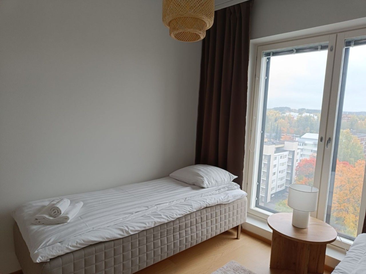 Une petite chambre avec un lit simple, donnant sur des immeubles et des arbres grâce à une grande fenêtre.