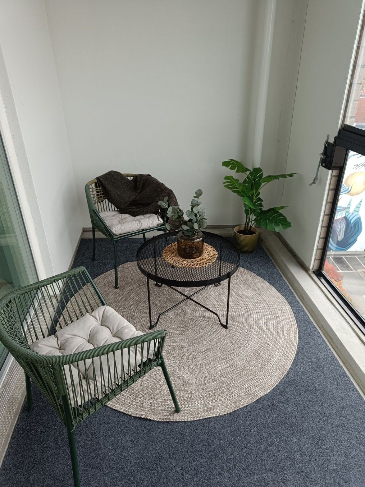 A balcony with a green chair, a small round table with a woven mat, and a potted plant.
