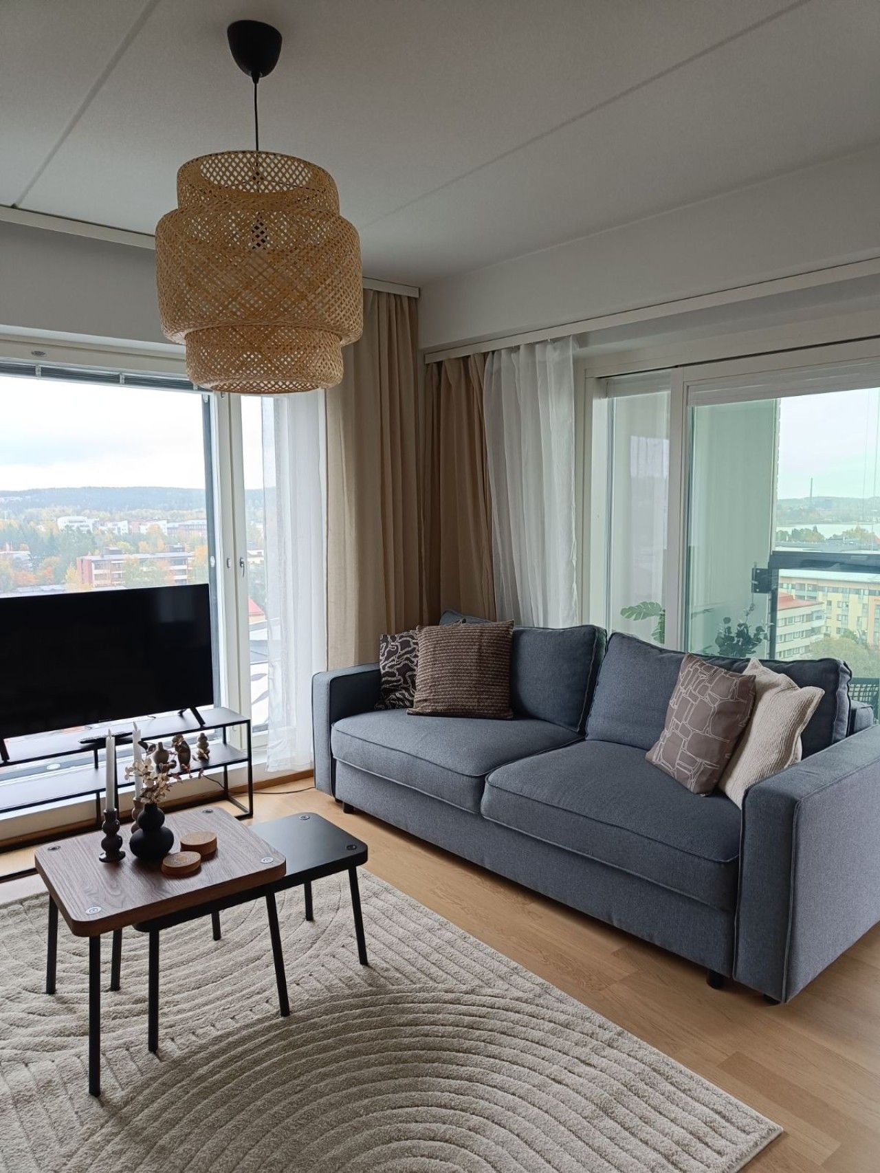 A modern living room with a grey sofa, a coffee table, a TV, and a large window with a view of buildings.