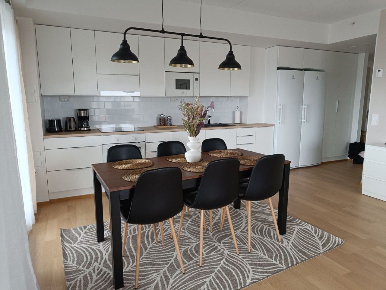Een moderne keuken met witte kastjes, een eettafel met zwarte stoelen en een kamerbreed tapijt met een patroon.