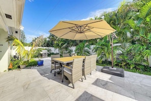 An outdoor patio with a table and chairs, a large umbrella, and lush tropical plants. - Beach in 5 Mins - Private Saltwater Pool BBQ (Deerfield Beach)