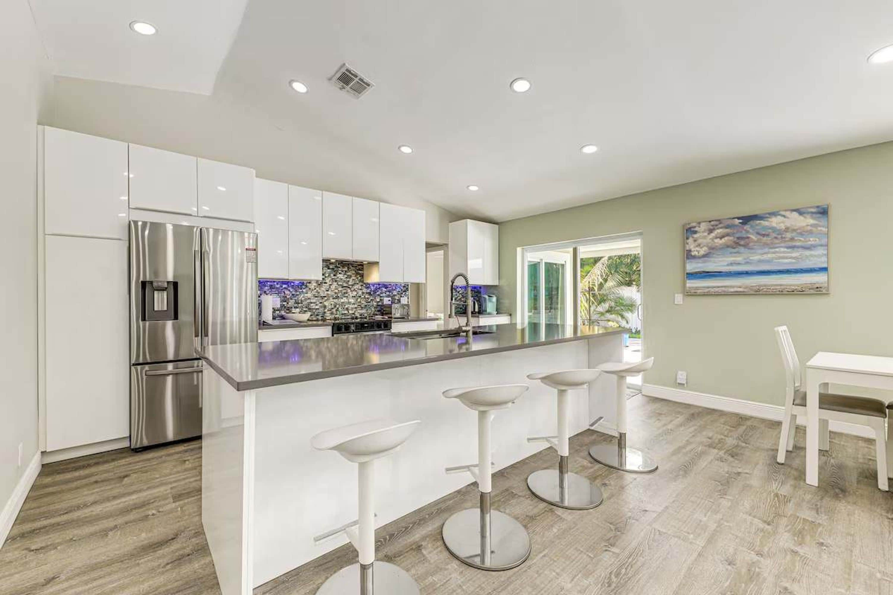 A modern kitchen with white cabinets, a stainless steel refrigerator, and a bar area with white stools.