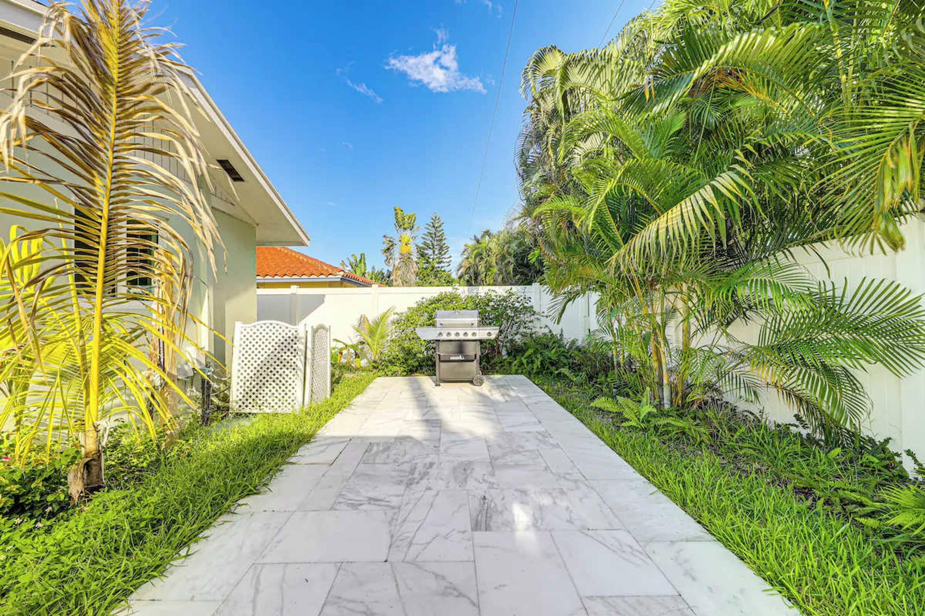A paved pathway leading to a barbecue grill, surrounded by lush greenery and a white fence.