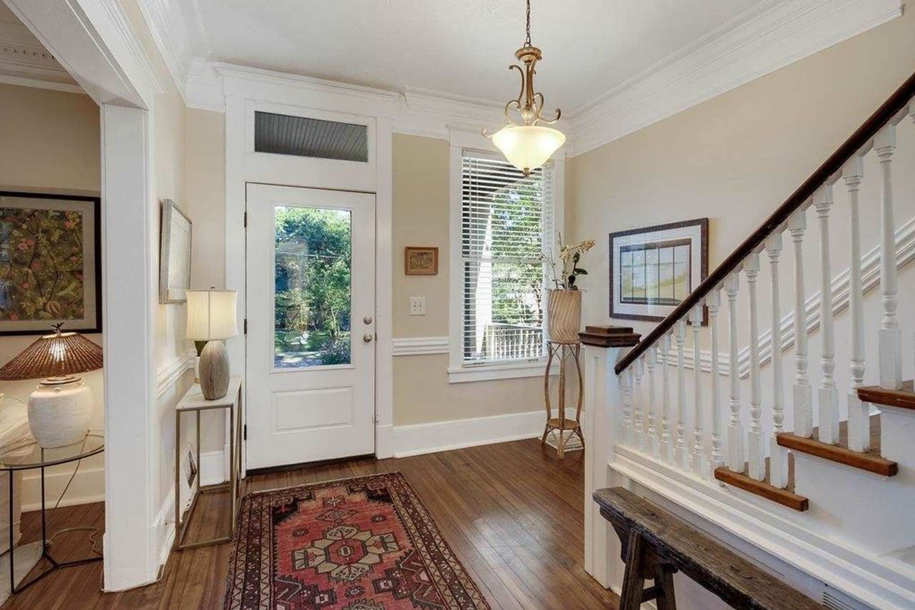 A hallway with a wooden floor, a patterned rug, a white door with a glass panel, a staircase with white balusters, and a hanging light fixture.