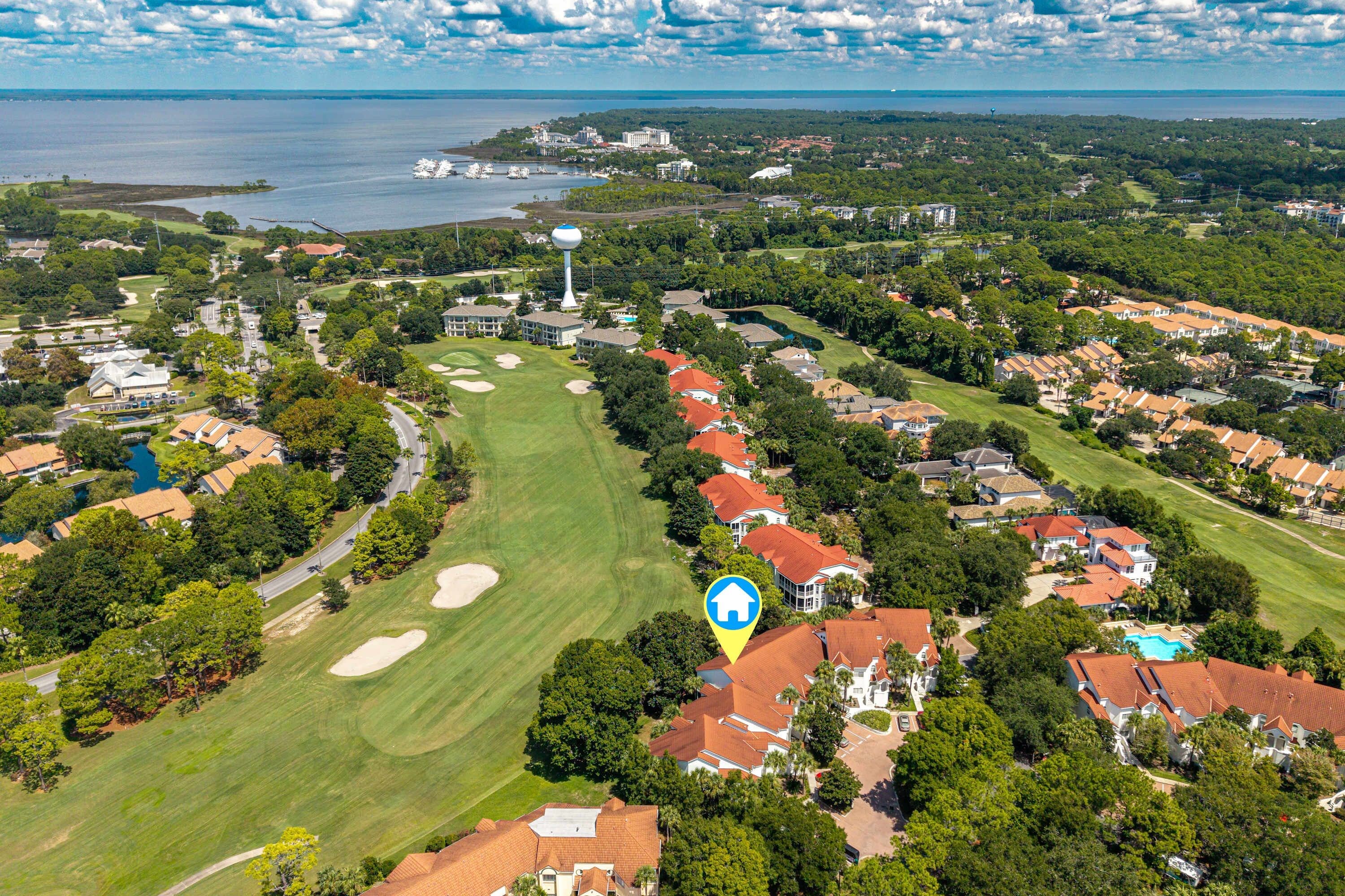 An aerial view of a golf course with a water tower, surrounded by residential areas and a body of water in the distance.
