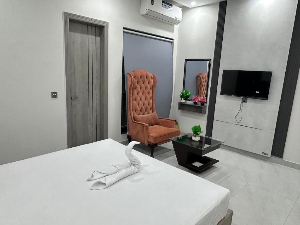 Habitación de hotel con una cama, un sillón tapizado, una mesita con una planta, un televisor de pantalla plana en la pared y una puerta.