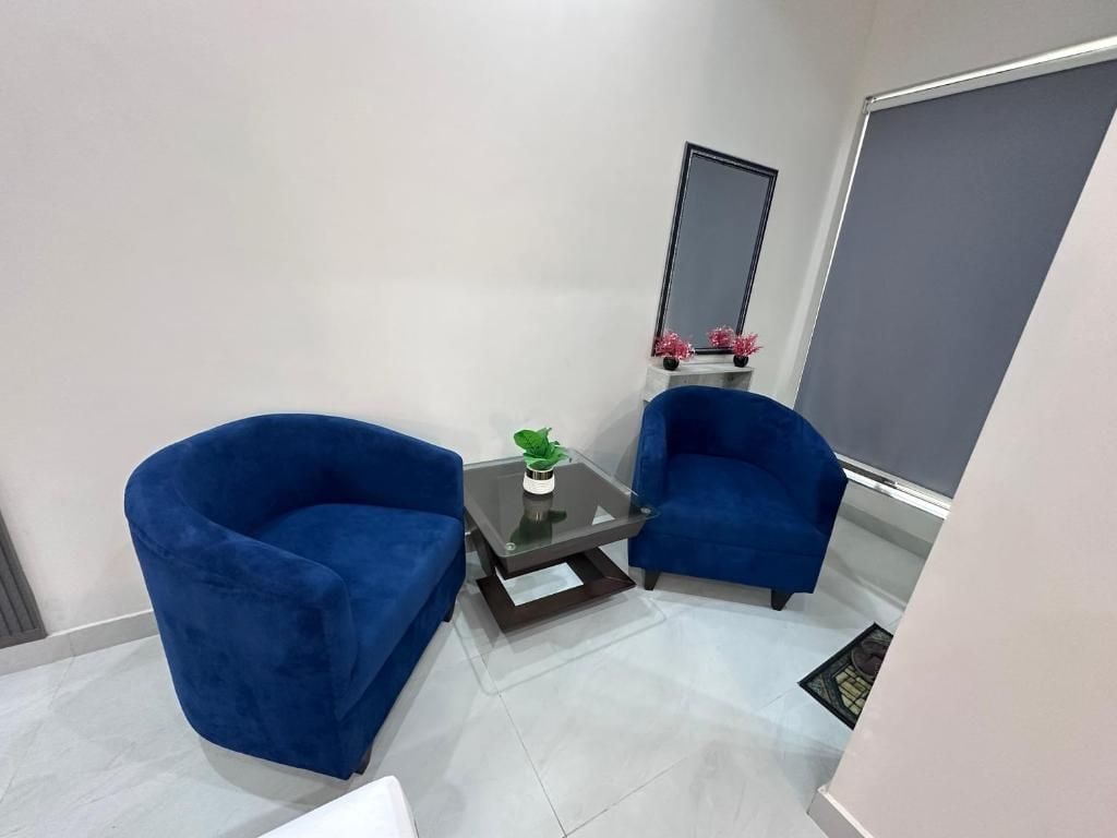 Ein kleines Zimmer mit zwei blauen Sesseln, einem Glastisch mit Pflanze, einem Spiegel und einem Fenster.
